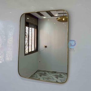 May include: Rectangular mirror with a gold-colored frame. The mirror is mounted on a white wall, reflecting a room with a window and patterned flooring. The mirror's rounded corners add a modern touch.