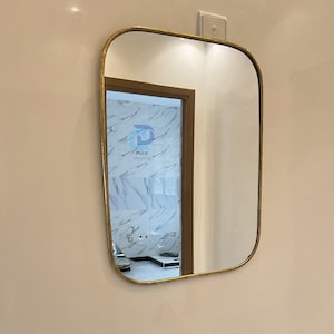 May include: A rectangular mirror with rounded corners and a gold-colored frame is mounted on a beige wall. The mirror reflects a glimpse of a bathroom with marble-like tiles and a door. A gold faucet is visible below.