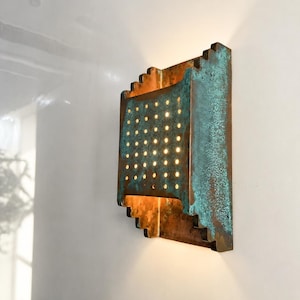 Könnte beinhalten: Eine rechteckige Wandleuchte mit einer verwitterten Kupferoberfläche und einer ausgeprägten türkisfarbenen Patina. Die Leuchte hat ein gestuftes Kantendesign und eine mittlere Platte mit einem Raster aus kleinen Löchern für warmes Licht.