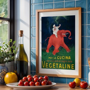 Könnte beinhalten: Ein gerahmtes Vintage-Werbeplakat für Vegetaline mit einem roten Elefanten, der von einem Koch geritten wird. Das Bild befindet sich in einer Küche mit einer Flasche Olivenöl, Tomaten und einer dekorativen Vase.
