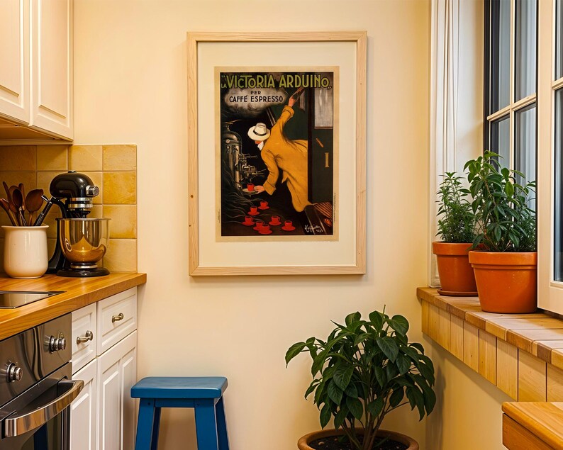 Puede incluir: Un p&oacute;ster vintage enmarcado de Victoria Arduino Caffe Espresso cuelga en una cocina. El p&oacute;ster presenta a un hombre con abrigo y sombrero amarillos, con tazas rojas. La cocina tiene gabinetes blancos, un taburete azul y plantas en macetas.