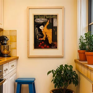 Puede incluir: Un p&oacute;ster vintage enmarcado de "Victoria Arduino Caffe Espresso" cuelga en una cocina. El p&oacute;ster presenta a un hombre con abrigo y sombrero amarillos. La cocina tiene armarios blancos, un taburete azul y plantas en macetas en un alf&eacute;izar.