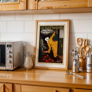 Puede incluir: Un p&oacute;ster vintage enmarcado de Victoria Arduino Caffe Espresso. La obra de arte presenta a un hombre con un abrigo amarillo sirviendo caf&eacute; espresso. La imagen est&aacute; sobre una encimera de madera con un microondas, utensilios y una jarra de metal.
