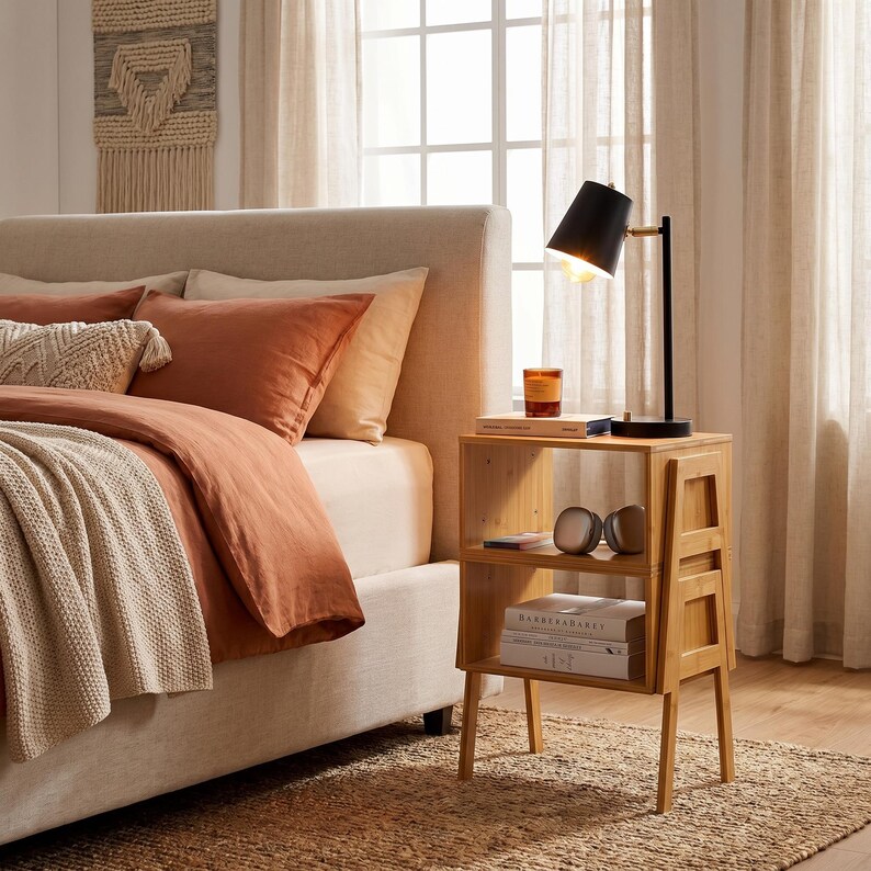 May include: A bedroom scene featuring a bed with rust-colored bedding and a woven throw. A small, light-colored wooden nightstand holds a black desk lamp, books, and decorative items. The room has neutral tones and natural light.