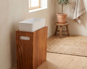 May include: A white rectangular storage bin sits atop a tall, wooden cabinet with a handle cutout. The cabinet has a rich, warm wood tone. The scene is set in a well-lit room with a small wooden stool, a potted plant, and a rug.