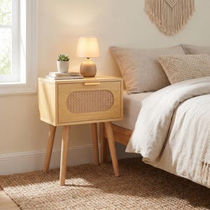 May include: A light wood nightstand with a woven panel on the drawer. A small lamp and a potted plant sit on top. The nightstand has four angled legs and a gold-colored handle.