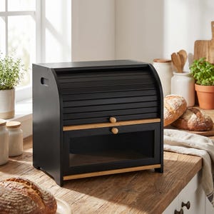 May include: A black wooden bread box with a roll-top lid and a clear glass window. The box has a natural wood trim and two wooden knobs. The bread box is sitting on a wooden countertop near a window.
