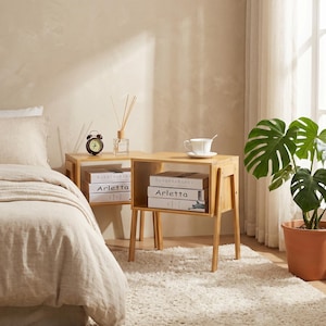 May include: Two small, light-colored wooden side tables with open storage compartments. Each table holds books, a small alarm clock, and a cup and saucer. A bed with a beige comforter is in the foreground, and a potted plant is in the background.