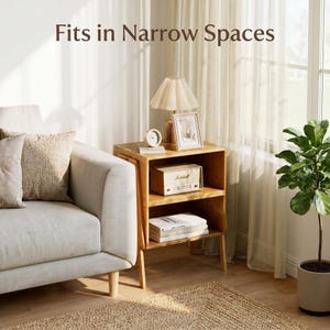 May include: A wooden side table with two shelves, holding a lamp, clock, picture frame, and a small radio. The text "Fits in Narrow Spaces" is at the top. The table is next to a sofa and a window with sheer curtains.