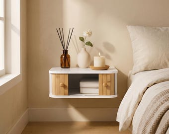 May include: A white wall-mounted floating shelf featuring two sliding bamboo doors with round wooden knobs. The shelf has an open center compartment containing two white books, and the top surface holds a reed diffuser, a vase, and a candle.