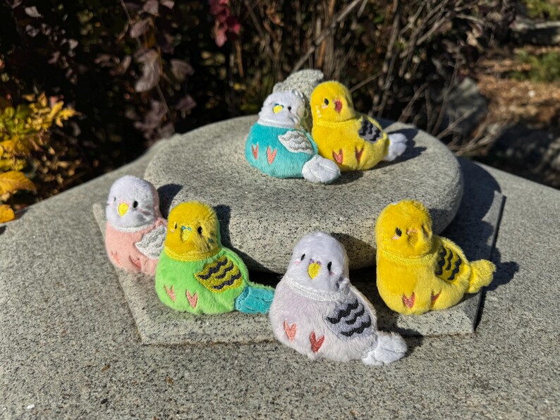 May include: A collection of plush budgie bird toys in various colors, including yellow, green, blue, pink, and white. The birds are arranged on a stone surface, with details like wings and beaks visible. The toys are soft and fluffy.