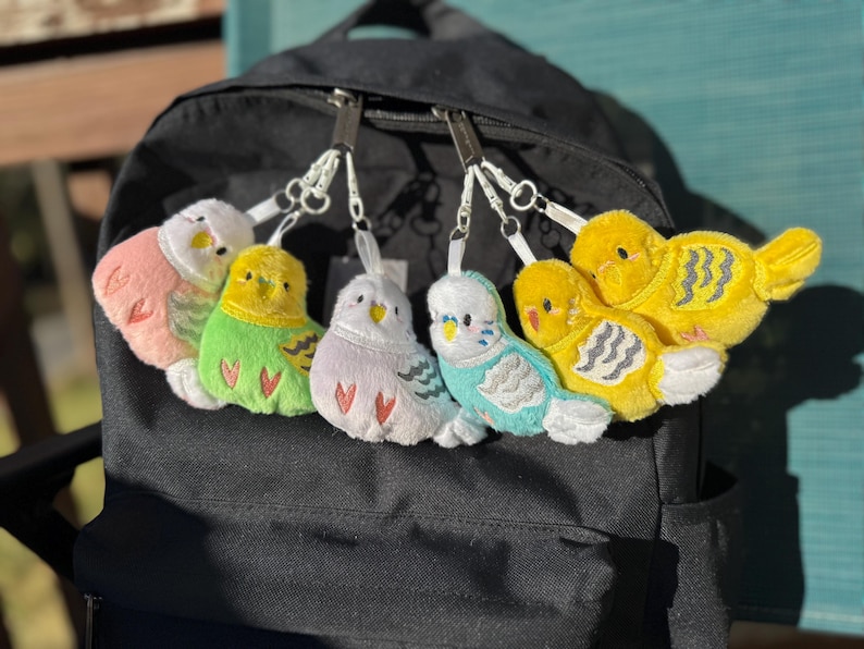 May include: A collection of six colorful plush bird keychains attached to a black backpack. The birds are in shades of pink, green, purple, blue, and yellow, with white accents and silver key rings. The birds have embroidered details.