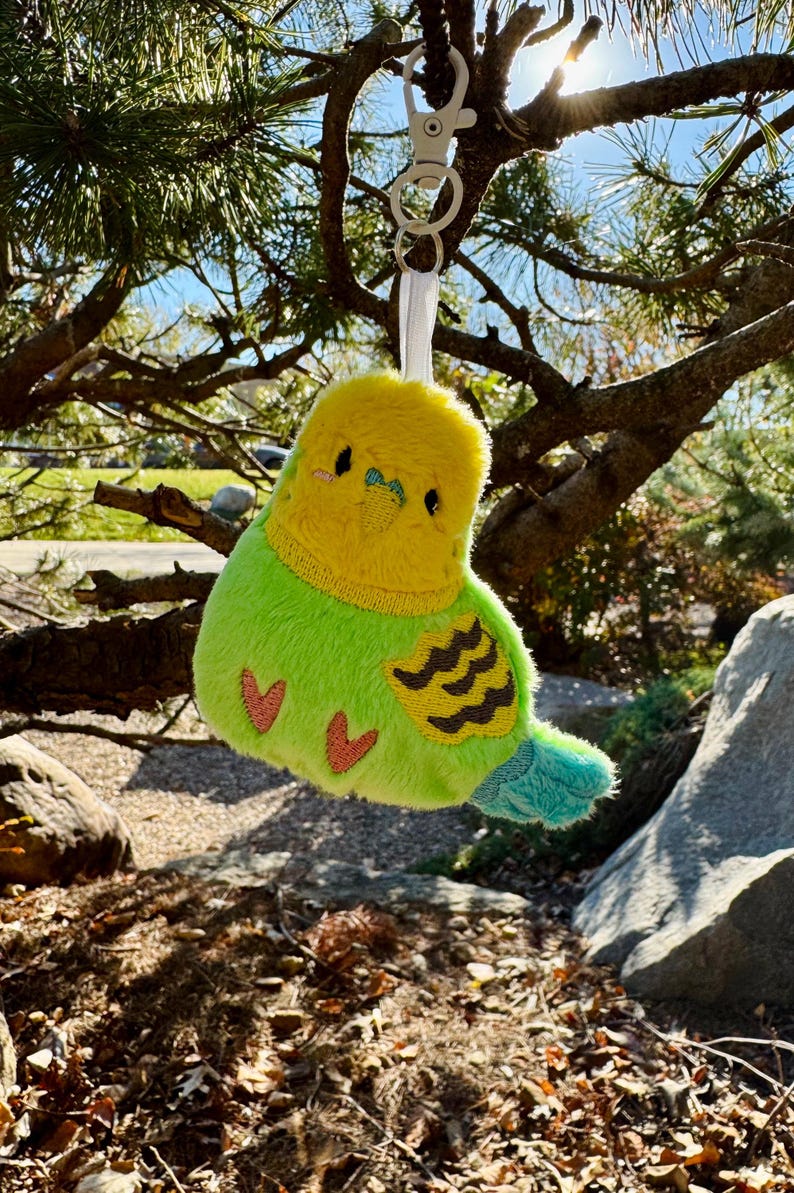 May include: A plush parakeet keychain in bright colors. The bird has a yellow head and chest, a green body, and a blue tail. It features embroidered details, including two small red hearts on the body. The keychain is attached to a silver clasp.