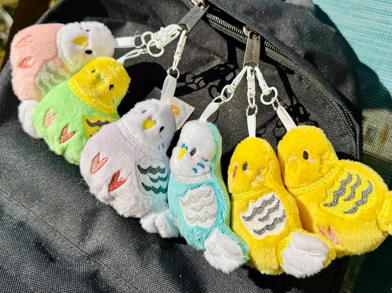May include: A collection of colorful plush bird keychains in various colors, including pink, green, purple, and yellow. Each keychain features a small, soft bird with embroidered details and a metal clasp for attachment.