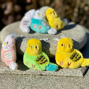 May include: A collection of plush parakeet keychains in various colors, including yellow, green, pink, and teal. The keychains are arranged on a stone surface, with some in the foreground and others in the background. Each keychain has a small metal loop.