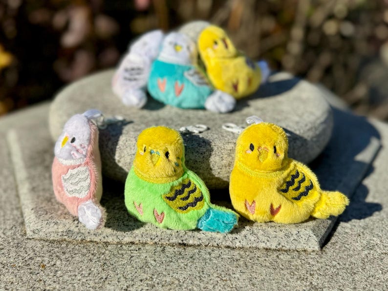 May include: A collection of plush parakeet keychains in various colors, including yellow, green, pink, and teal. The keychains are arranged on a stone surface, with some in the foreground and others in the background. Each keychain has a small metal loop.