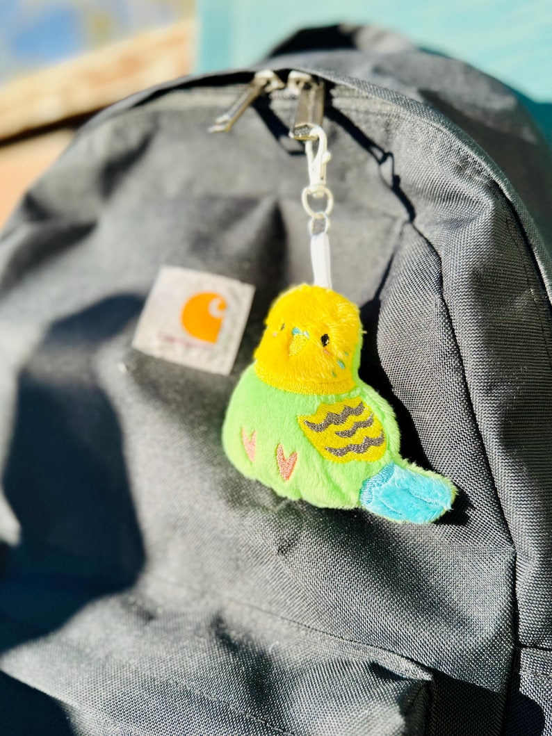 May include: A plush bird keychain with a yellow head, green body, and blue tail hangs from a backpack. The bird has black wing markings and pink heart details. The backpack is gray with a Carhartt logo.