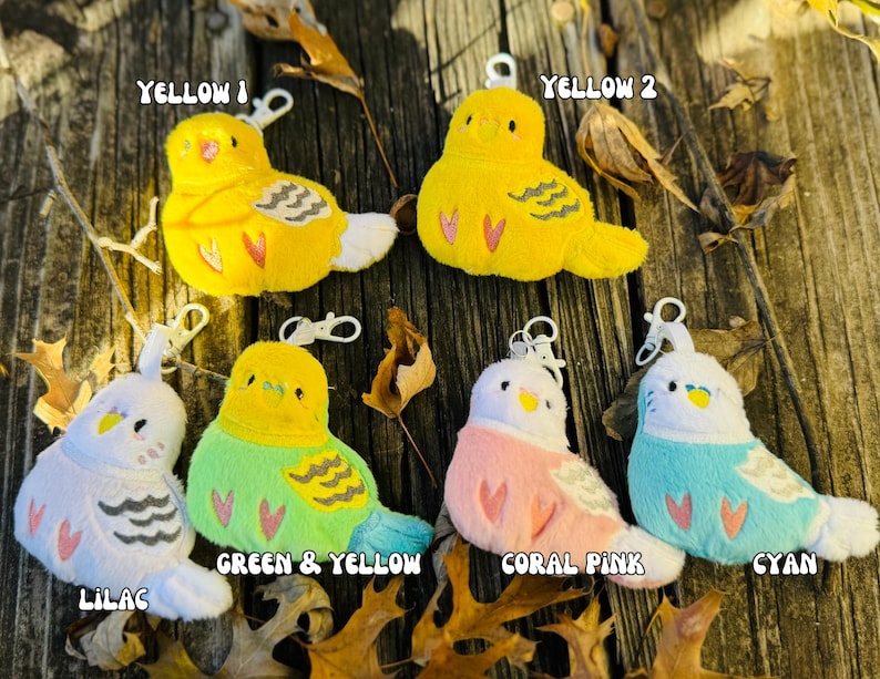 May include: A collection of plush bird keychains in various colors, including yellow, lilac, green, coral pink, and cyan. Each bird has a white belly, a small beak, and a keychain attachment. The text "Yellow 1", "Yellow 2", "Green & Yellow", "Coral Pink", "Cyan", and "Lilac" are visible.