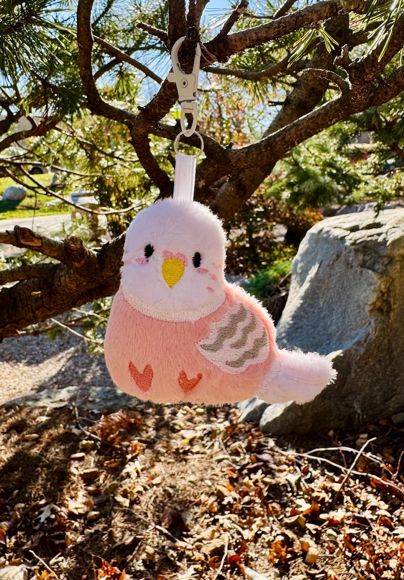 May include: A plush pink and white bird-shaped keychain with a silver clasp, hanging from a tree branch. The bird has a yellow beak, black eyes, and gray wing details. The keychain is a cute accessory.