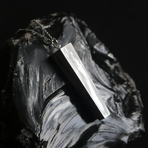 May include: A silver pendant necklace with a rectangular, brushed metal design. The pendant is hanging on a black rock.