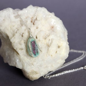 May include: A silver chain necklace with a green and pink gemstone pendant. The pendant is wrapped in silver wire and has a natural, irregular shape.
