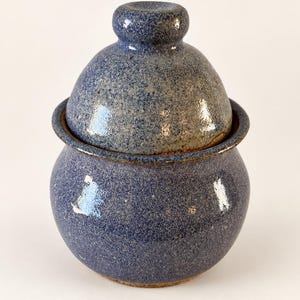 May include: A round ceramic jar with a matching lid, featuring a speckled blue glaze over a textured surface. The container has a bulbous body and a small knob handle on top of the lid, with a visible brown clay rim where the lid meets the base.