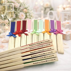 Peut inclure: Une collection d'éventails en bois avec des tissus colorés en bleu, rouge, bordeaux, rose, vert, vert clair, orange, violet et magenta. Chaque éventail présente un petit nœud en ruban et le texte gravé MARTA Y PEDRO 8-8-2020.