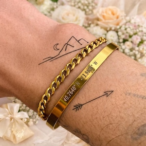 May include: A gold-toned metal curb chain bracelet and a smooth gold-toned metal cuff bracelet engraved with the coordinates 40.7648 N -73.9808 W. The bracelets are worn on a wrist featuring black ink tattoos of a mountain range with a crescent moon and an arrow.