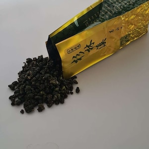 May include: A gold and green foil pouch of loose leaf tea with Chinese characters on the front. The pouch is open and tea leaves are spilling out onto a white surface.