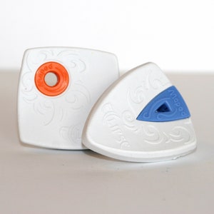 May include: Two white plastic fabric softener sheets with a decorative design. One sheet has a round orange button with the word "Nappe" printed on it. The other sheet has a blue triangular button with the word "Nappe" printed on it.