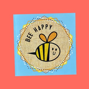May include: A square card with a light blue background featuring a central tan burlap-textured hexagon. Inside the hexagon is a cartoon bee with yellow and black stripes and the text BEE HAPPY printed in black capital letters above the bee.