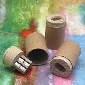 May include: A wooden pencil sharpener with two blades and a natural wood finish, next to three cardboard tubes. The tubes are cylindrical and made of brown paper. The background is a colorful abstract painting.