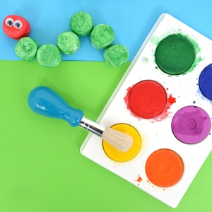 May include: A set of watercolors with a blue paintbrush and a green caterpillar made of round objects. The colors in the watercolor set are green, red, yellow, purple, and orange.