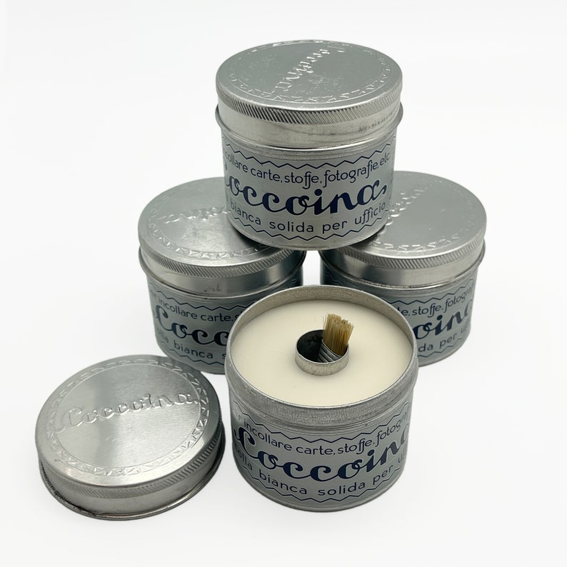 May include: A stack of four silver tins with white glue inside. The tins have a blue label with the text "incollare carte, stoffe, fotografie etc. "occoina" bianca solida per ufficio."
