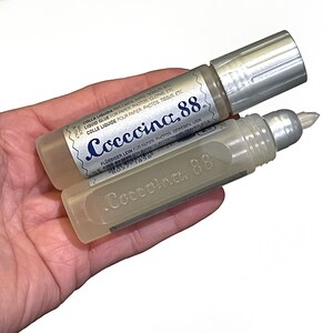 May include: Two clear plastic bottles of Coccoina 88 liquid glue. The bottles have silver caps and are labeled in multiple languages. The text on the label reads "Coccoina 88" in blue lettering.