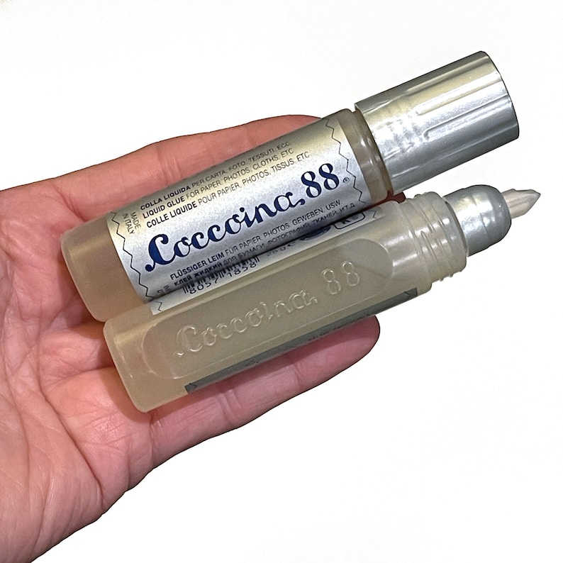May include: Two clear plastic bottles of Coccoina 88 liquid glue. The bottles have silver caps and are labeled in multiple languages. The text on the label reads "Coccoina 88" in blue lettering.
