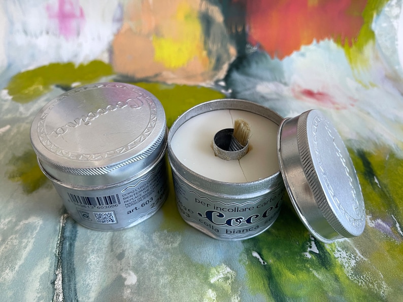 May include: Two silver tins of white glue with a brush attached to the lid. The tin on the right is open and the glue is visible. The text on the tin reads "per incollare carte e tessuti colla bianca"
