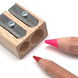 May include: A wooden pencil sharpener with two blades and the text "Made in Germany". Two colored pencils, one with a pink tip and the other with a red tip, are positioned near the sharpener. The sharpener has the brand name "KUM" on it.