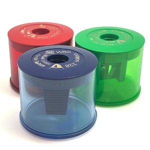 May include: Three colorful pencil sharpeners in red, green, and blue. Each sharpener has a cylindrical design with a transparent lower section and a solid-colored top. The top of the blue sharpener has the text "Wax Crayon 226" and "Runde Spitze".