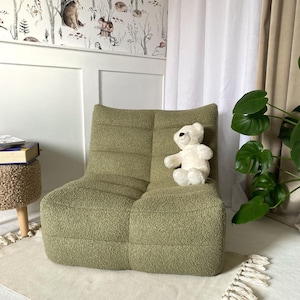 Könnte beinhalten: Ein hellgrüner, strukturierter Sessel mit einem weißen Teddybären. Der Stuhl hat ein modernes Design und steht auf einem weißen Teppich. Der Hintergrund umfasst eine Wand mit Waldtapete und einen weißen Vorhang.
