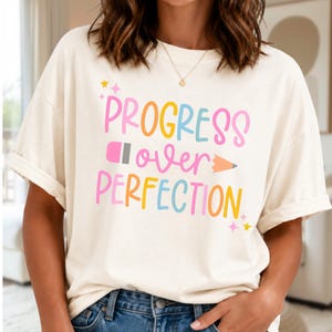 May include: A cream-colored short-sleeve t-shirt featuring the colorful text "PROGRESS over PERFECTION" with a pencil graphic integrated into the word "over". The design includes small star accents in pink and yellow, printed on the front of the shirt.