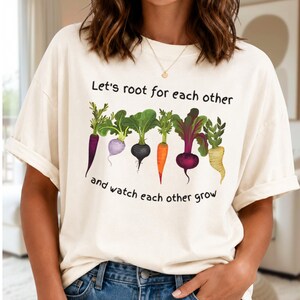 Puede incluir: Una camiseta de manga corta de color crema con un gráfico de seis coloridas hortalizas de raíz en fila. Sobre las verduras aparece el texto "Let's root for each other" y debajo el texto "and watch each other grow" en letra negra.