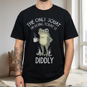 Könnte beinhalten: Ein schwarzes T-Shirt mit kurzen Ärmeln und einer Grafik eines grünen Frosches, der eine dampfende Tasse hält. Der Text über dem Frosch lautet THE ONLY SQUAT I'M DOING TODAY IS und darunter steht DIDDLY in weißen Großbuchstaben.