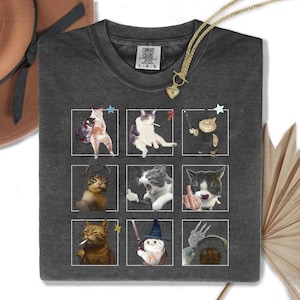 May include: Dark gray Comfort Colors t-shirt featuring a grid of nine cat illustrations in white squares. The cats are depicted in various poses, some with accessories. A gold necklace with a heart pendant is draped over the shirt.