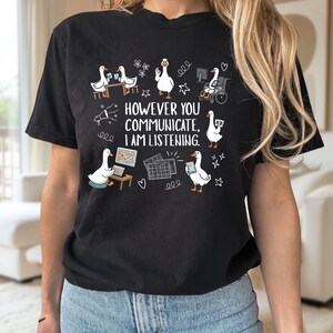 Puede incluir: Una camiseta negra de manga corta con un gráfico de gansos blancos usando varios métodos de comunicación. El texto dice "HOWEVER YOU COMMUNICATE, I AM LISTENING". El diseño incluye ilustraciones de gansos con lenguaje de señas, una silla de ruedas y tabletas.