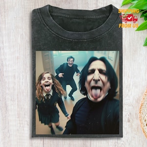 Könnte beinhalten: Dunkelgraues T-Shirt mit einem Selfie-ähnlichen Bild von drei Personen, die ihre Zungen herausstrecken. Das Bild zeigt eine junge Frau in Schuluniform und zwei Männer. Das Shirt hat einen Vintage-Look.