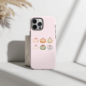 May include: A light pink phone case featuring a collection of watercolor pumpkins. The pumpkins are in various colors, including pink, orange, green, and white, each adorned with a delicate pink bow. The case has a black camera module and black side buttons.