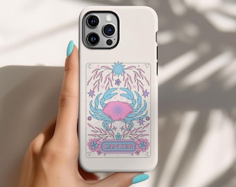 Puede incluir: Carcasa blanca para iPhone con una ilustración rosa y azul de un cangrejo y la palabra "Cáncer" en un círculo rosa. La ilustración está rodeada de estrellas y flores.