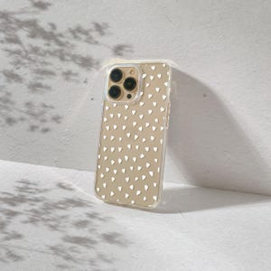 Peut inclure: Une coque de téléphone transparente avec un motif de cœurs blancs. La coque est conçue pour un iPhone avec un renflement de caméra.