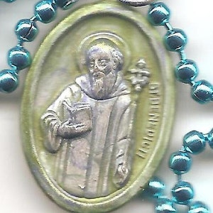 May include: A green oval-shaped metal pendant with a silver image of a saint holding a book and a staff. The pendant is on a blue beaded chain.